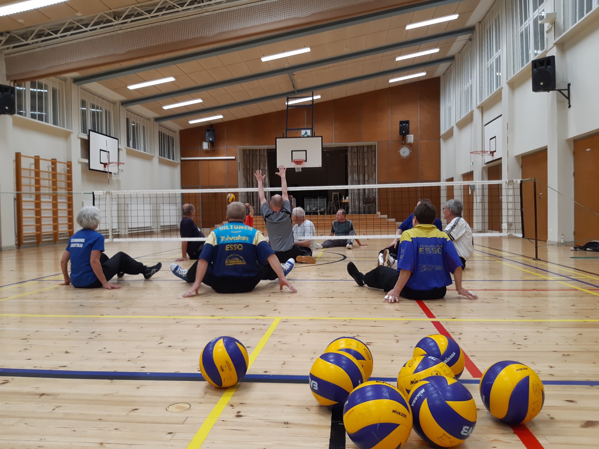 Joukko ihmisiä pelaamassa istumalentopalloa liikuntasalissa, etualalla sinikeltaisia lentopalloja.